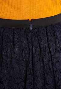 Jupe en dentelle noire avec un motif floral texturé, une taille ajustée avec un détail de fermeture éclair, associée à un haut orange à côtes.