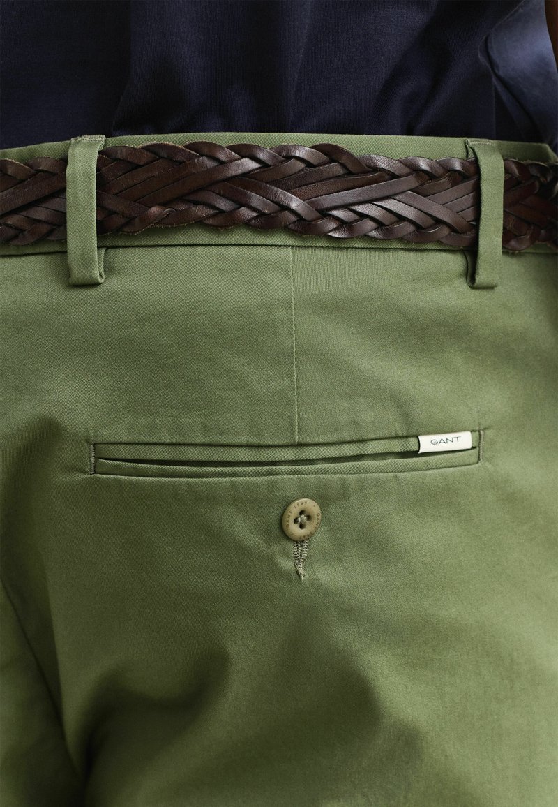 Primer plano de pantalones verde oliva con un bolsillo trasero abotonado y un cinturón trenzado de cuero marrón oscuro sujetando una camisa azul marino.