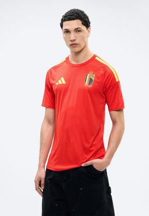 Joven vestido con una camiseta roja de fútbol de Bélgica con rayas y logotipos amarillos, de pie con una mano en el bolsillo sobre fondo blanco.