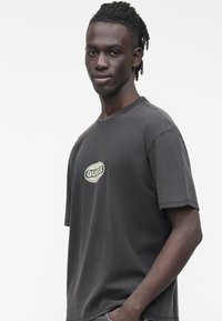 Sivé bavlnené tričko s voľným strihom. Na ľavej hornej časti hrude má kruhové zeleno-biele logo s nápisom „GUESS“.