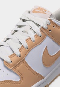 Λευκό και μπεζ αθλητικό παπούτσι Nike με λευκά κορδόνια, μπεζ λογότυπο Nike swoosh και μπεζ ραμμένες επιφάνειες στα πλάγια και την περιοχή των δαχτύλων.