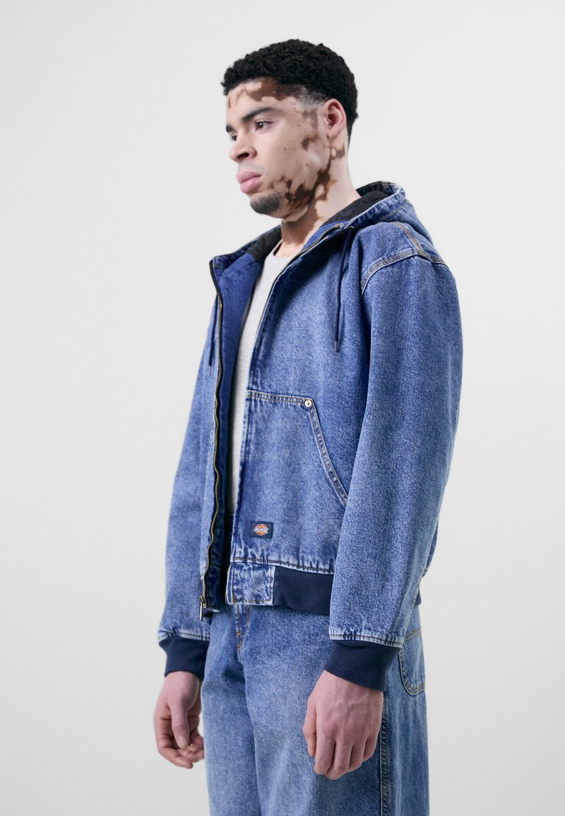 Giacca di jeans in blu chiaro, con cappuccio, chiusura con zip, tasche laterali e polsini a costine blu scuro. Indossata con pantaloni in denim coordinati.