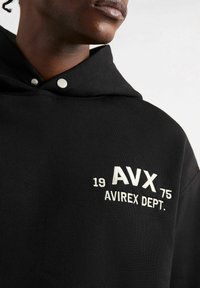 Felpa nera con cappuccio in cotone, caratterizzata da ricami color argento con la scritta "AVX 1975 AVIREX DEPT." Due dettagli con bottoni a pressione sul colletto.