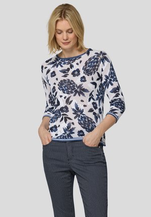 Frau mit blondem Haar, die ein weißes und marineblau geblümtes Langarmshirt und marineblaue Nadelstreifenhose mit Händen in den Taschen trägt.
