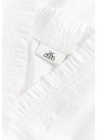 Îmbrăcăminte albă cu un design plisat pe decolteu și accente ondulate. Include o etichetă din material cu detaliu logo negru.