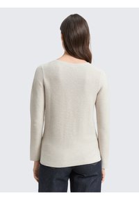 Femme aux longs cheveux bruns portant un pull côtelé à manches longues beige clair et un jean bleu foncé, vue de dos sur un fond uni.