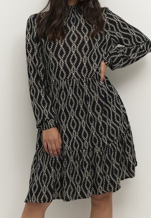 Femme portant une robe noire à manches longues avec un motif de feuilles beige ondulées, une main sur la hanche, l'autre reposant à ses côtés.