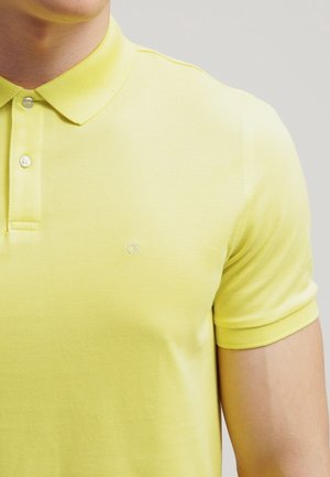 Gros plan d'une personne portant un polo à manches courtes jaune clair avec un petit logo "ck" sur la poitrine et deux boutons au col.