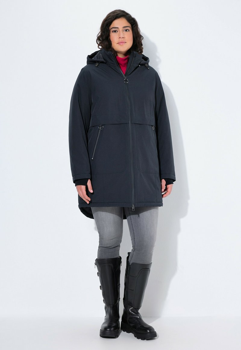 Černá voděodolná parka s kapucí, předním zipovým zapínáním, dvěma bočními zipovými kapsami a delším zadním dílem. Nosená přes červený rolák a šedé džíny.