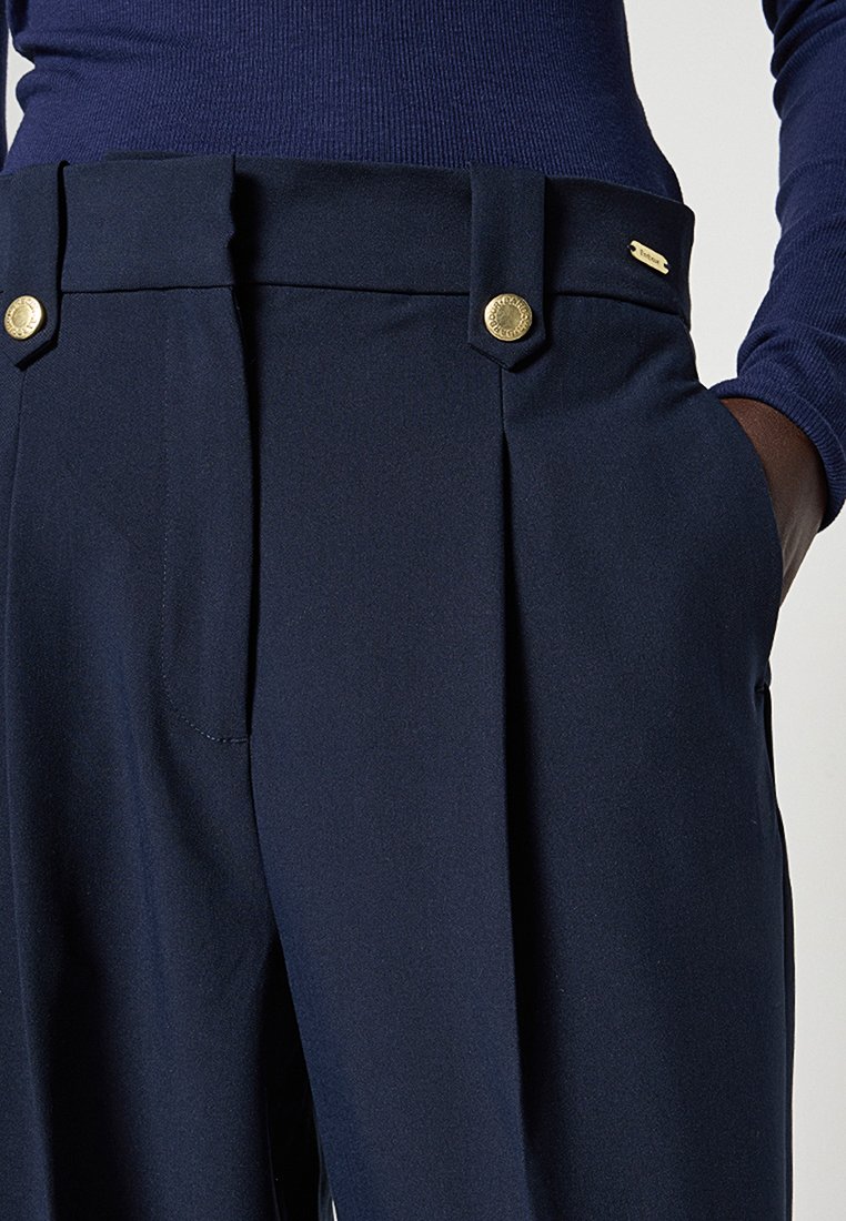 Pantalon taille haute bleu marine avec plis et détails de boutons dorés, porté avec un haut à manches longues côtelé bleu marine, main dans la poche.