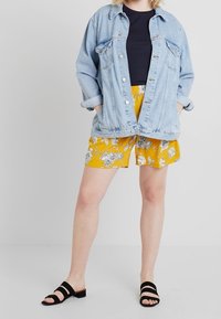 Džínová bunda, černé tričko a žluté květinové šortky. Šortky mají volný střih a elastický pas. Celkový look doplňují černé sandály na pásky.