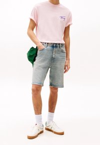 Hombre con camiseta rosa de mangas enrolladas, pantalones cortos de mezclilla claros, calcetines blancos, deportivas blancas y sosteniendo una gorra verde detrás de su espalda.