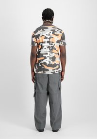Šedé maskáčové tričko s oranžovými akcenty a velkým bílým nápisem "ALPHA INDUSTRIES", spárované se šedými cargo kalhotami s bočními kapsami.