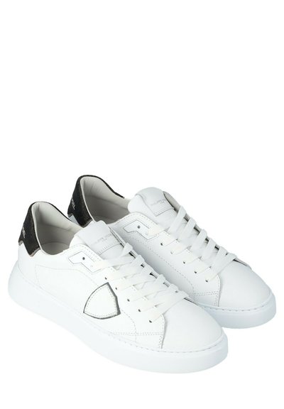 Un paio di sneaker bianche basse con dettagli neri sul tallone, design con lacci e un distintivo patch triangolare cucito sul lato.