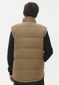 Eine Person trägt eine beigefarbene gepolsterte Weste über einem schwarzen Langarmshirt, von hinten betrachtet vor einem schlichten weißen Hintergrund.