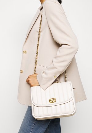 Žena oblečená v béžovém saku a modrých džínách nese na rameni bílou prošívanou kabelku se zlatým řetízkovým popruhem a zapínáním.