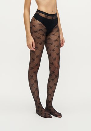 Femme portant des collants noirs transparents à motif répétitif, debout devant un fond clair uni.