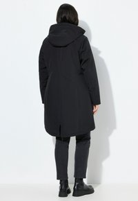 Femme se tenant de dos, portant un manteau à capuche noir arrivant aux cuisses, un jean noir et des bottines noires, sur fond blanc.
