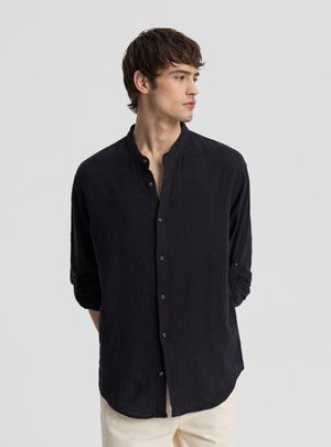 Giovane uomo che indossa una camicia nera a maniche lunghe con colletto alla mandarina, guarda verso destra su uno sfondo grigio uniforme.