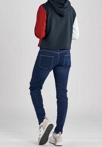 Obcisły, skrócony sweatshirt z kapturem w granatowe, czerwone i białe rękawy, zestawiony z niebieskimi, wąskimi dżinsami i białymi trampkami z szarymi podeszwami.