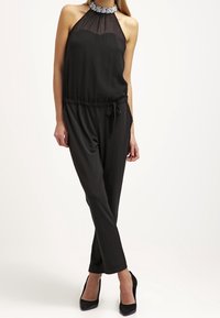 Mono negro sin mangas con escote transparente, cintura ceñida y piernas ajustadas. Presenta un collar decorativo y una tela suave.
