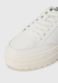 Witte sneaker met een glad leren bovenwerk, platte veters en een dikke, gestructureerde rubberen zool met een geribbeld patroon.