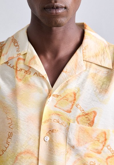 Chemise à manches courtes avec boutons dans des nuances de jaune clair et d'orange, dotée d'un motif subtil avec du texte et d'une poche poitrine. Tissu léger.