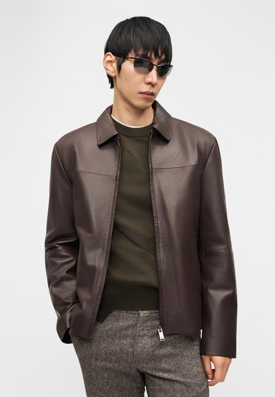 Άνδρας που φοράει σκούρο καφέ δερμάτινο μπουφάν, πουλόβερ ελιάς, γκρι υφασμάτινο παντελόνι και σκούρα γυαλιά ηλίου, στέκεται μπροστά σε απλό φόντο.