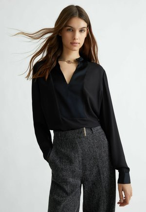 Blusa di seta nera con scollo a V, caratterizzata da una toppa testurizzata e polsini a contrasto. Abbinata a pantaloni a vita alta grigi a fantasia.