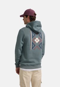 Sudadera con capucha en verde azulado tenue, con un diseño de patrones geométricos en azul marino, rojo y crema en la parte posterior. Mezcla de algodón, textura suave.