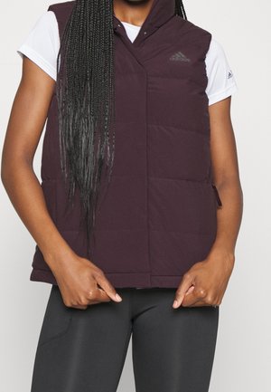 Personne avec de longs cheveux tressés portant un gilet matelassé Adidas violet par-dessus un t-shirt blanc à manches courtes et des leggings noirs, les mains tenant l'ourlet du gilet.