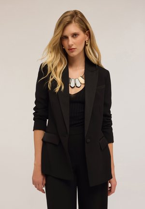 Black tailored blazer with textured fabric, single button closure, and two front pockets. Worn over a black layered top and paired with a chunky necklace.
