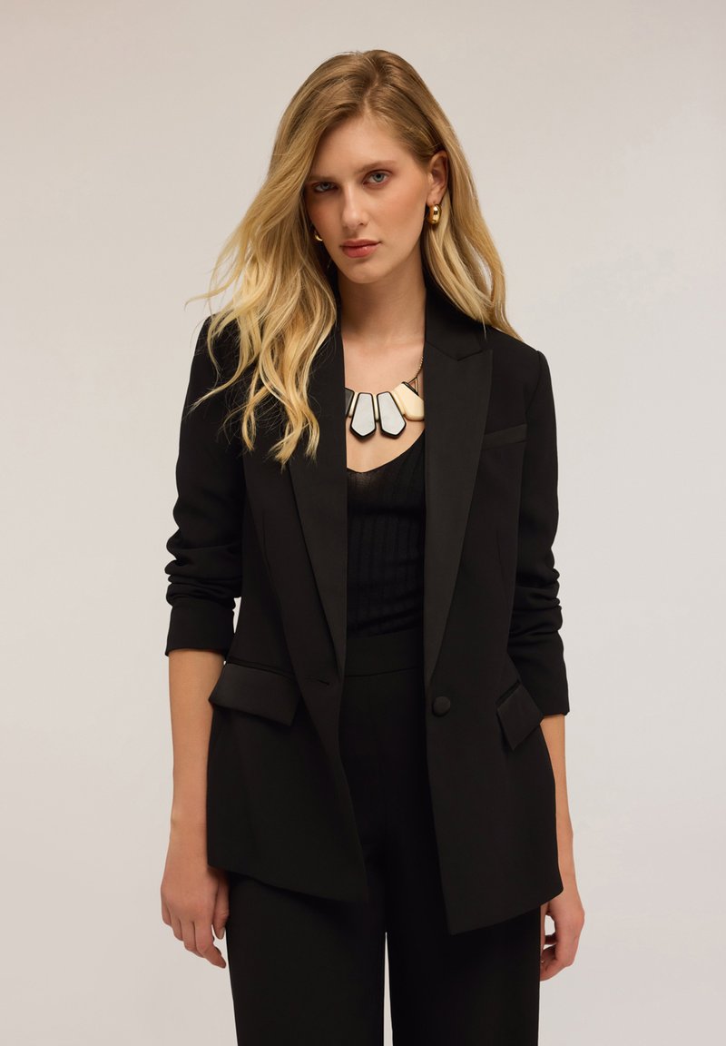 Blazer nero su misura con tessuto texturizzato, chiusura a un bottone e due tasche frontali. Indossato sopra un top nero a strati e abbinato a una collana chunky.