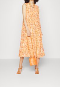 Orange floralmønstret ærmeløs kjole med lagdelt design. Kombineret med en flettet orange håndtaske og stroppede hæle i matchende tone.