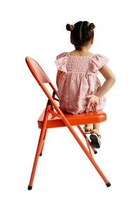 Robe rose brodée avec des manches volantées sur une enfant assise sur une chaise pliante orange vif. La chaise est dotée d'un cadre en métal robuste.