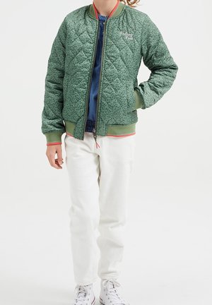 Barn iklädd en grönt mönstrad quiltad jacka med texten "Feeling Fine", vita byxor och vita sneakers, stående med ena handen i fickan.