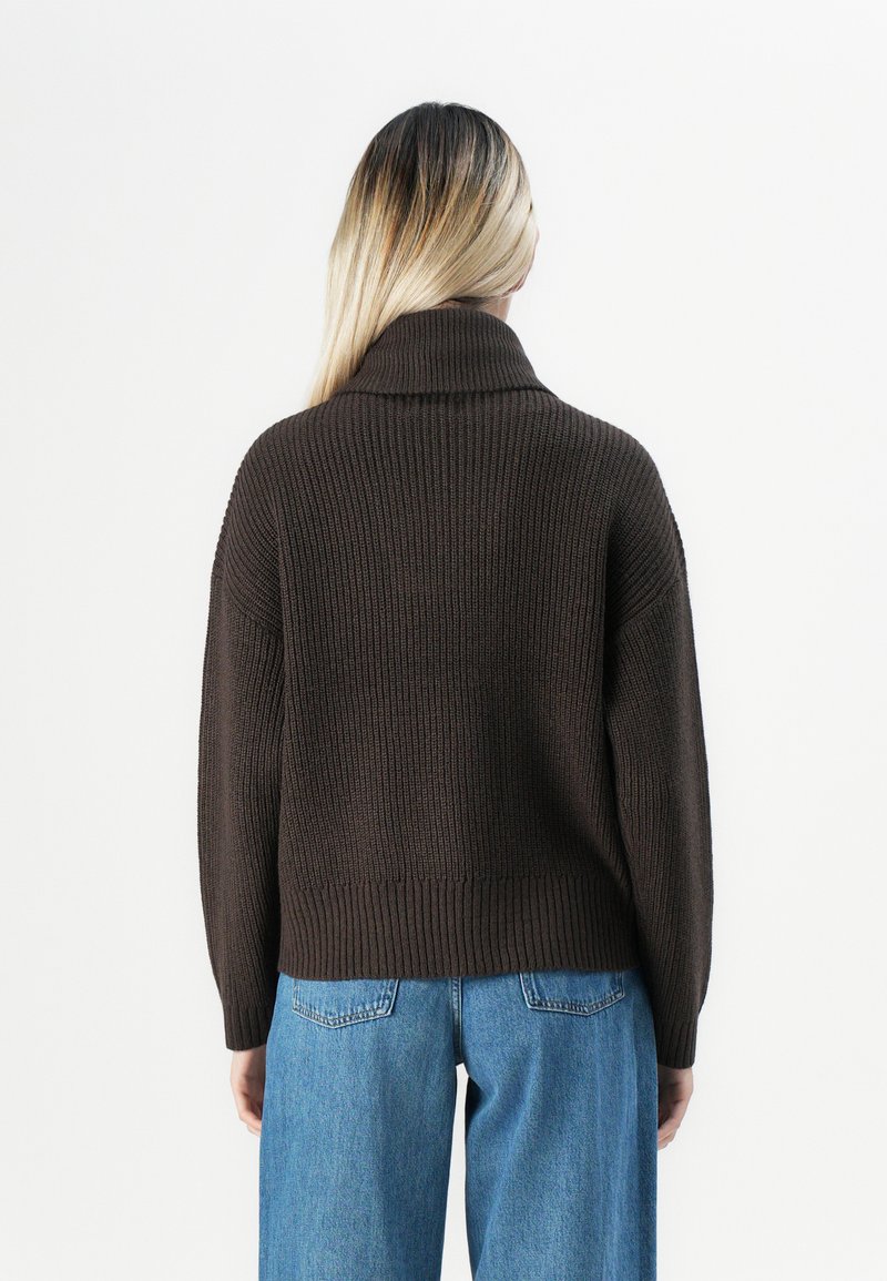 Personne avec de longs cheveux blonds portant un pull en col roulé côtelé marron foncé et un jean en denim bleu, se tenant de dos.