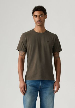 Mężczyzna z krótkimi kręconymi włosami, ubrany w oliwkowo zielony T-shirt i niebieskie dżinsy, stojący na jednolitym jasnoszarym tle.