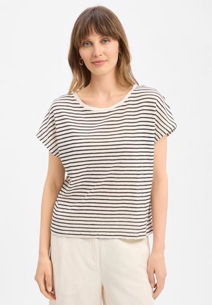 Frau trägt ein weißes Kurzarmshirt mit dünnen schwarzen horizontalen Streifen und cremefarbenen Hosen, steht vor einem einfarbigen Hintergrund.