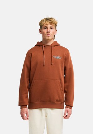 Giovane uomo con capelli biondi indossa una felpa marrone Billabong con tasca frontale e pantaloni color crema, in piedi davanti a uno sfondo semplice.