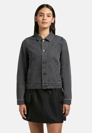 Kobieta ubrana w zapinaną na guziki ciemnoszarą kurtkę dżinsową z długimi rękawami oraz czarną spódnicę z przednimi kieszeniami, stojąca na jednolitym tle.