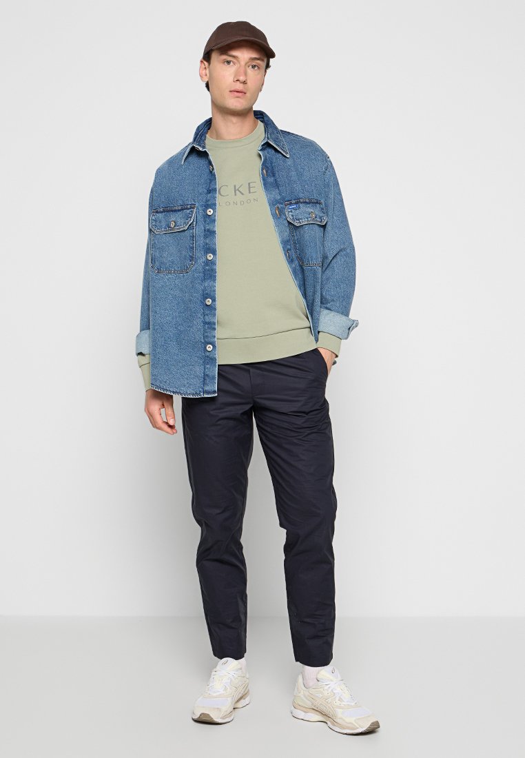 Jeune homme portant une casquette marron, une veste en denim bleue sur un sweat-shirt vert clair, un pantalon noir et des baskets blanches, se tenant contre un mur clair.