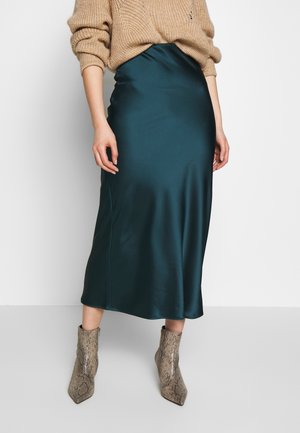 Person trägt einen tealfarbenen Satin-Midirock, einen beigen grob gestrickten Pullover und knöchelhohe Stiefel mit Schlangenledermuster vor weißem Hintergrund.