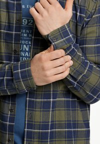 Camisa de cuadros en verde y azul marino con un patrón de rejilla blanca, que cuenta con puños abotonados y un corte relajado. Superpuesta sobre una camiseta gráfica azul.