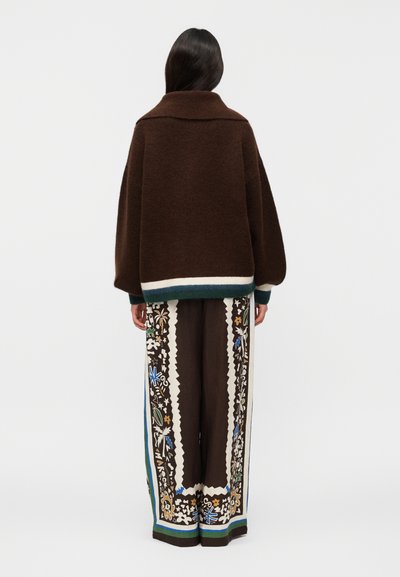 Γυναίκα με μακριά σκούρα μαλλιά φοράει φαρδιά καφέ πουλόβερ και καφέ παντελόνια με φαρδιά μπατζάκια με πολύχρωμα λουλουδάτα σχέδια, απεικονίζεται από πίσω.