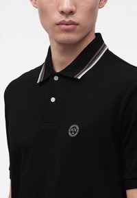 Homme portant un polo noir avec un col rayé et un écusson rond avec un logo sur la poitrine.