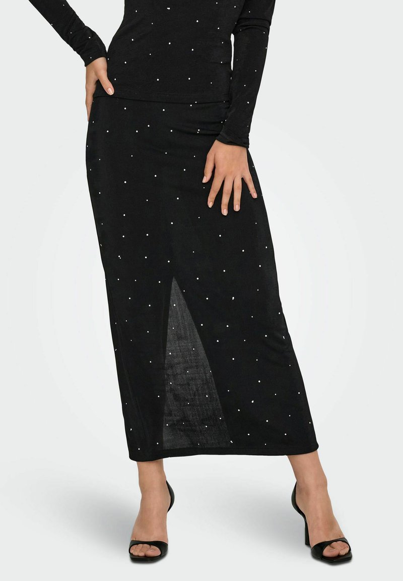 Falda midi negra con una abertura frontal, adornada con pequeñas tachuelas plateadas. Hecha de una tela suave y texturizada. Combinada con zapatos de tacón negros.