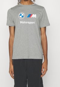 Šedé bavlněné tričko s logem BMW Motorsport a bílým textem, s krátkými rukávy a kulatým výstřihem. Černé šortky viditelné dole.