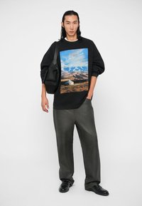 Uomo che indossa una felpa nera con stampa di paesaggio montano, pantaloni larghi grigi, scarpe nere e porta una borsa a spalla nera.