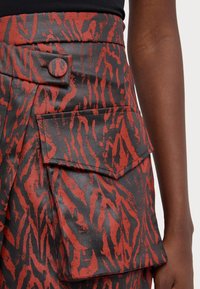 Pantalons cargo à rayures tigre noirs et rouges avec un bouton à l'avant, de grandes poches latérales et une texture brillante. Gros plan sur la taille et le détail des poches.